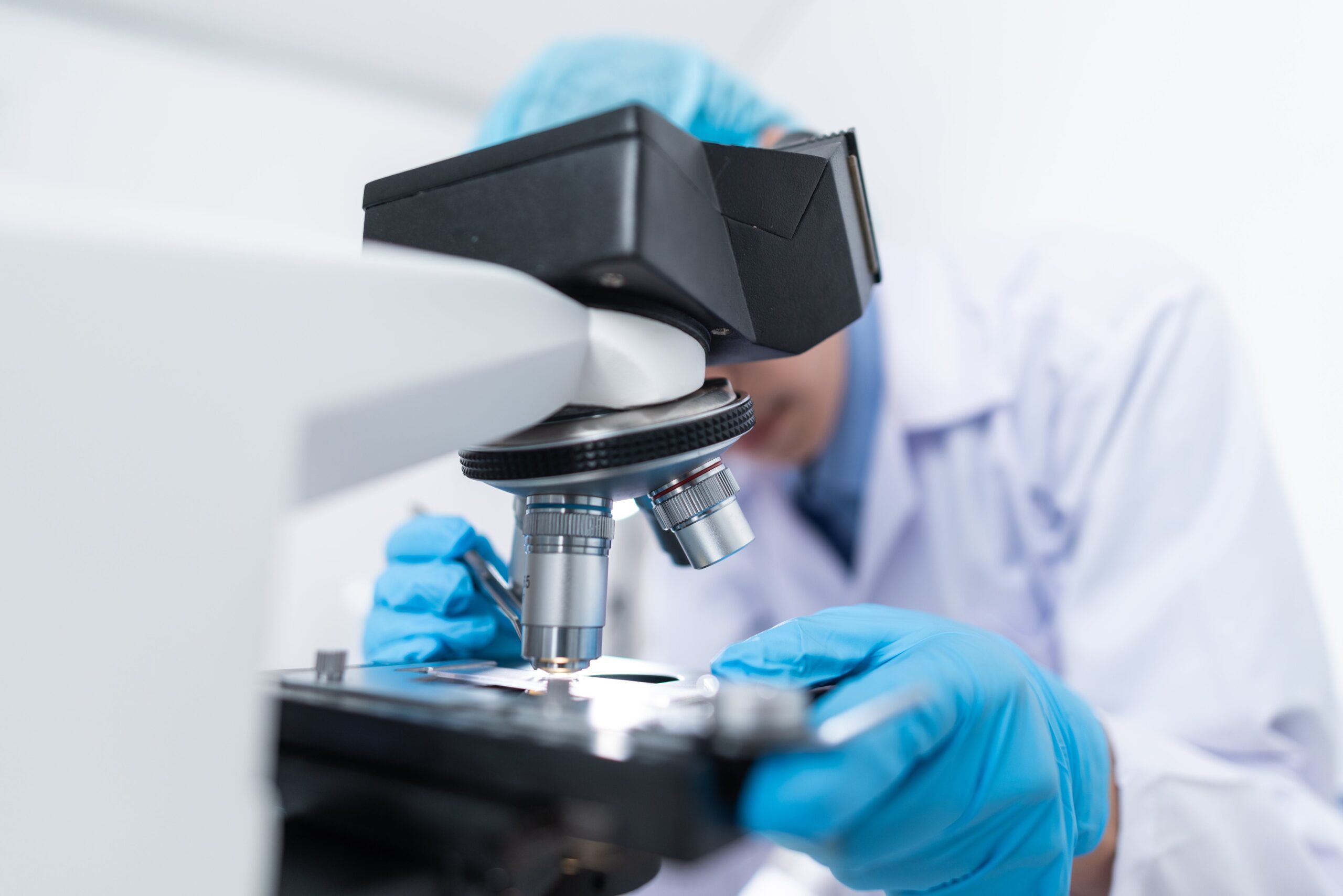 A laboratory worker wearing a white coat and rubber gloves using a microscope to embody the research undertaken.