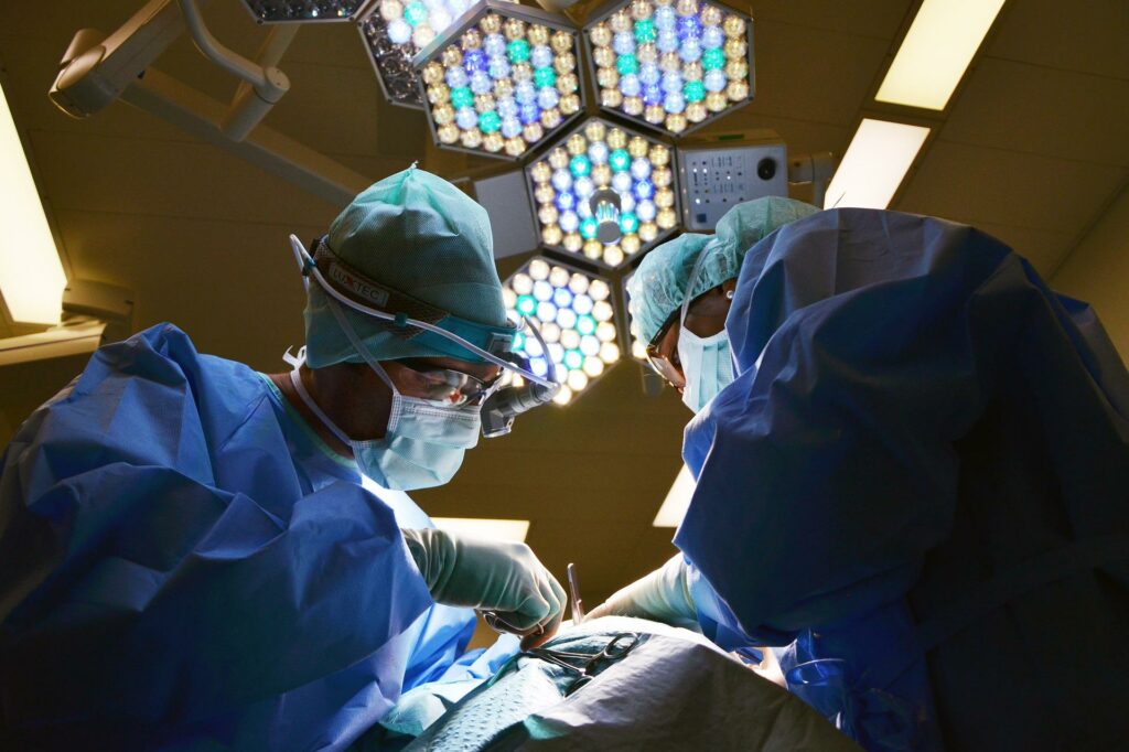 Two surgeons wearing PPE operating on a patient with lights on behind them